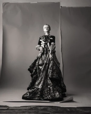 Full-length shot of a fair-skinned fashion model in an elaborate floral gown with a large bow, posing against a gray backdrop.