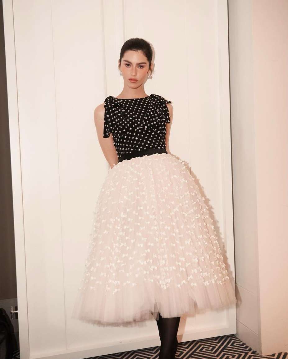 Elegant woman in a polka dot top and white tulle skirt, posing indoors with fair lighting against a white wall and geometric carpet.