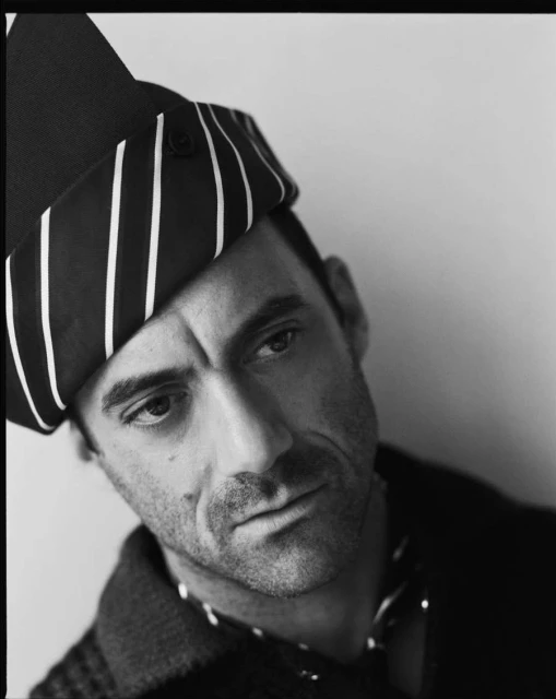 Black and white portrait of an adult male with a striped head wrap, posing indoors against a neutral background with soft, diffused lighting.
