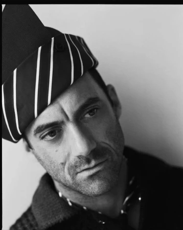 Black and white portrait of an adult male with a striped head wrap, posing indoors against a neutral background with soft, diffused lighting.