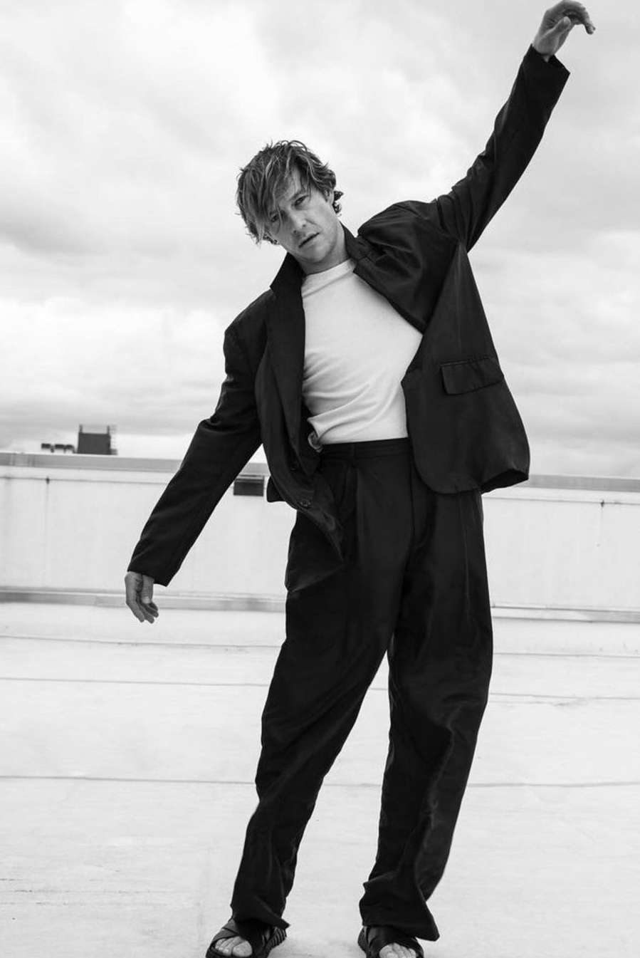 Man wearing a dark oversized suit jacket, white t-shirt, and dark trousers standing on a rooftop against an overcast sky