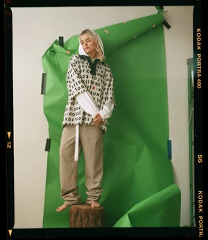 Woman standing barefoot on a tree stump against a green seamless paper backdrop wearing a patterned tunic and beige trousers