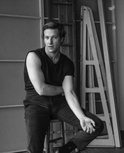 Black and white portrait of an adult man sitting on a stool with his arms crossed in front of windows and architectural elements.