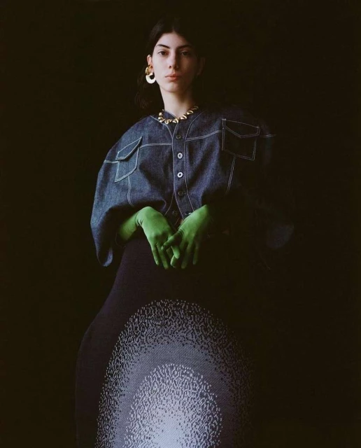 A young woman poses in a denim jacket, vibrant green gloves, and a patterned skirt against a dark backdrop, highlighted by studio lighting.