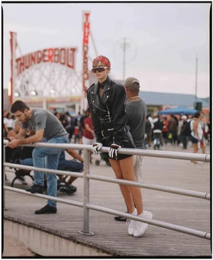 A woman in a leather jacket poses on a boardwalk in front of the Thunderbolt rollercoaster with a crowd of people in the background.