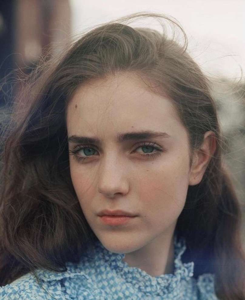 Close-up portrait of a young woman with long brown hair wearing a blue gingham patterned top against an outdoor background.