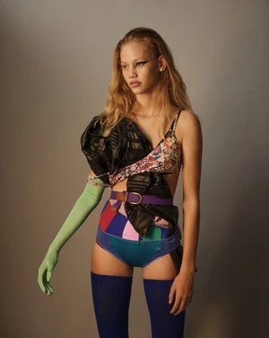 A young woman poses in a colorful patchwork outfit against a neutral backdrop, showcasing avant-garde fashion and style in a studio setting.