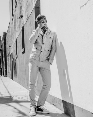 Monochrome image of a blond man in a utility jacket leaning against a white wall outdoors on a sunny day.