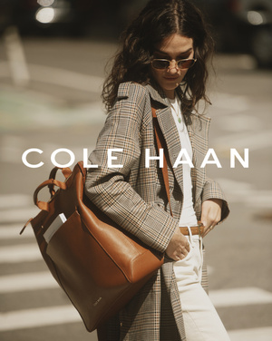 Fashionable woman in a brown plaid coat and sunglasses holds a brown leather backpack in an outdoor urban setting on a sunny day.
