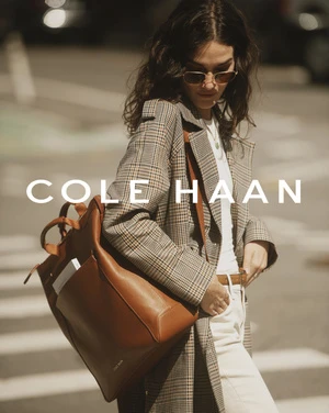 Fashionable woman in a brown plaid coat and sunglasses holds a brown leather backpack in an outdoor urban setting on a sunny day.