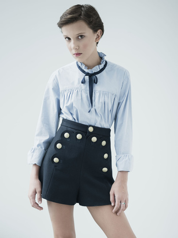 Young woman with short brown hair wearing a light blue blouse and dark blue shorts with white buttons poses in a photo studio.