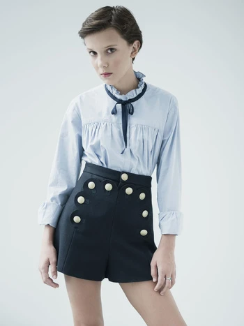 Young woman with short brown hair wearing a light blue blouse and dark blue shorts with white buttons poses in a photo studio.