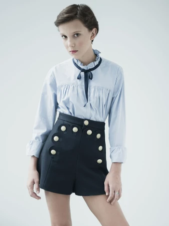 Young woman with short brown hair wearing a light blue blouse and dark blue shorts with white buttons poses in a photo studio.