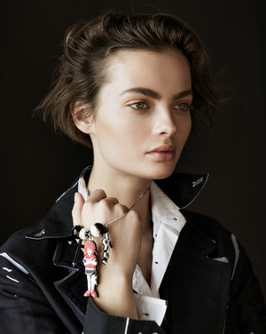Close-up portrait of a woman with short brown hair, wearing a black and white jacket, holding a doll necklace against a dark background.