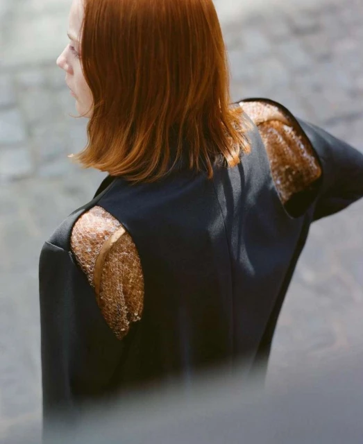 Back view of a woman with red hair in a black jacket featuring shoulder cutouts with mesh fabric, on a neutral outdoor background.