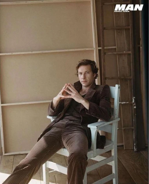 A man in a brown checkered suit sits on a white chair, looking at the camera with his hands clasped together in a light-filled indoor space.