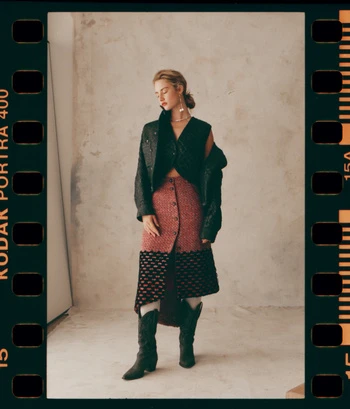 Woman poses indoors, wearing a dark cropped jacket, printed skirt with western boots, against a textured backdrop in a film frame.