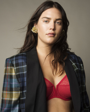 Portrait of a brunette adult woman wearing a plaid blazer, red bra, and gold earrings against a gray background in a studio setting.