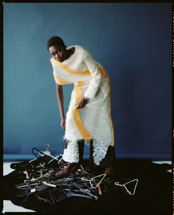 Adult woman in a yellow-and-white dress stands next to pile of black clothes hangers against a blue backdrop in a studio.