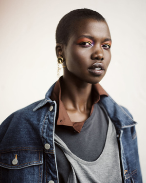 Close-up portrait of a Black woman with short hair wearing a denim jacket, colorful eye makeup, and gold earrings in a studio setting.
