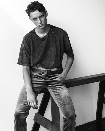 Monochrome portrait of a young male model seated on a wooden structure wearing a dark t-shirt and belted trousers.