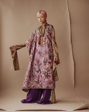 A pink-haired model poses in a studio wearing a floral-print kimono-style coat over purple wide-leg pants against a beige background.