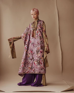 A pink-haired model poses in a studio wearing a floral-print kimono-style coat over purple wide-leg pants against a beige background.