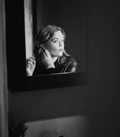 Black and white portrait of a woman adjusting an earring while looking into a framed wall mirror.
