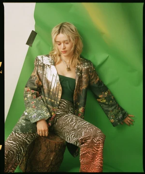 Blonde woman poses in a patterned green blazer and pants, seated on a log against a green studio backdrop with subtle yellow light.