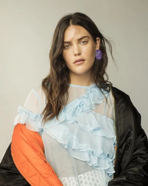 Portrait of a woman wearing a sheer blue ruffled top with a black and orange jacket draped over her shoulders.