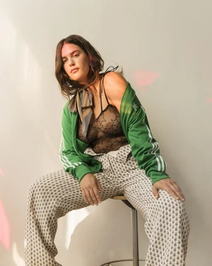 Woman sitting on a stool wearing a green track jacket and patterned trousers against a plain wall.