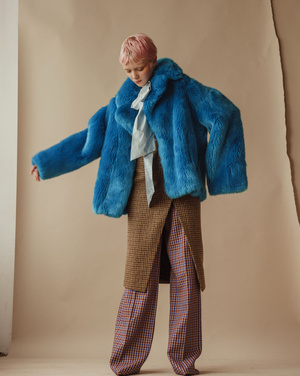 A young person with pink hair poses in a photo studio wearing a blue faux fur coat, pale blue top, and plaid patterned skirt and pants.