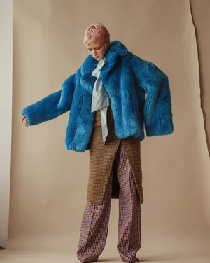 A young person with pink hair poses in a photo studio wearing a blue faux fur coat, pale blue top, and plaid patterned skirt and pants.