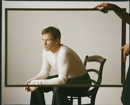 Adult man in a white long-sleeve shirt seated in a dark wood chair, framed by a dark rectangular border held by two dark-skinned hands.