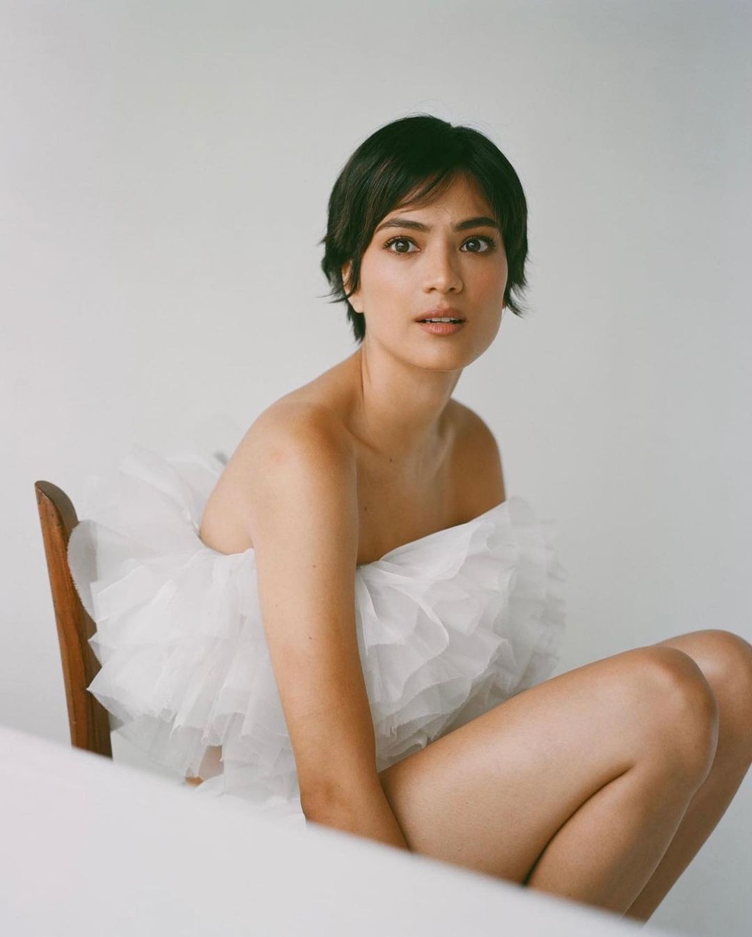 Portrait of a woman with short dark hair wearing a white ruffled top seated on a wooden chair against a plain white backdrop.