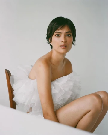 Portrait of a woman with short dark hair wearing a white ruffled top seated on a wooden chair against a plain white backdrop.
