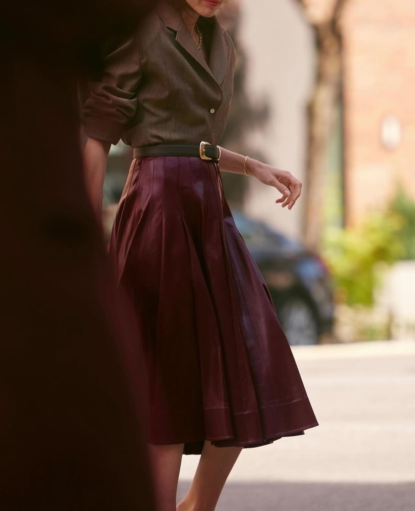 Fashionable woman in brown jacket and burgundy skirt walking outdoors, showcasing a chic and sophisticated style in natural light.