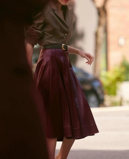 Fashionable woman in brown jacket and burgundy skirt walking outdoors, showcasing a chic and sophisticated style in natural light.