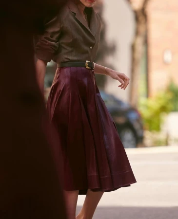 Fashionable woman in brown jacket and burgundy skirt walking outdoors, showcasing a chic and sophisticated style in natural light.