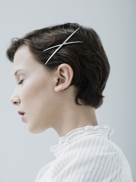 Profile portrait of a fair-skinned woman with short brown hair wearing a white ruffled blouse and metal hair clips against a light grey background.