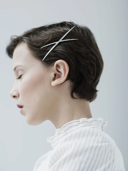 Profile portrait of a fair-skinned woman with short brown hair wearing a white ruffled blouse and metal hair clips against a light grey background.
