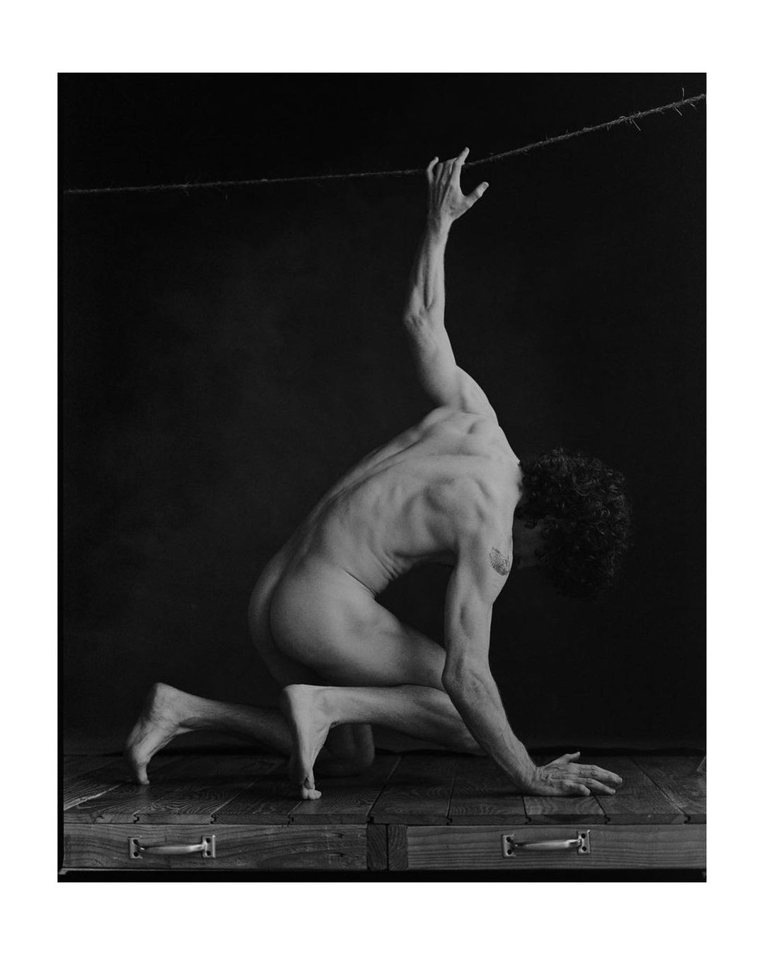 A nude adult male figure kneeling, reaching up to a branch with thorns in a black and white studio shot with wooden drawers.