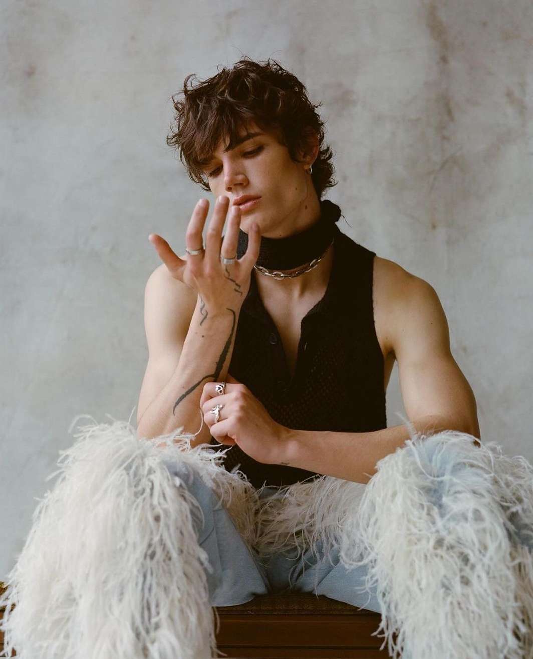 Young man with dark curly hair wearing a black sleeveless top and feather-trimmed trousers posing indoors