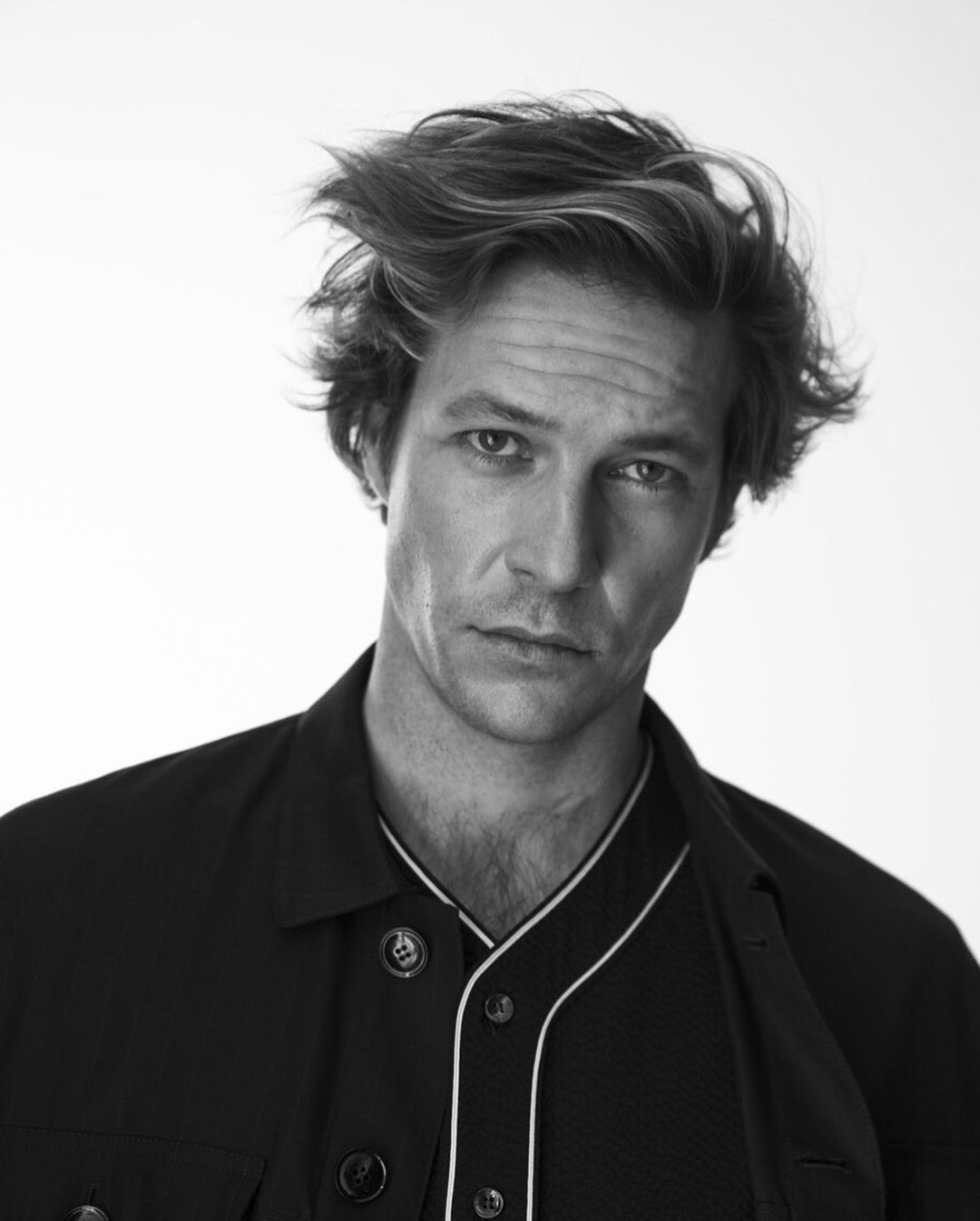 Black and white portrait of a handsome man with messy hair wearing a dark button-down shirt against a bright white background.