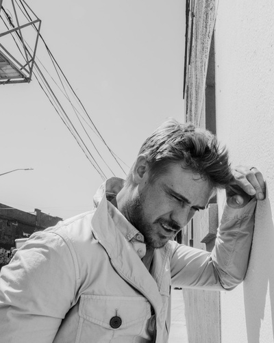 Monochrome portrait of adult male in a collared jacket, head tilted, hand in hair, with an urban background of building and utility lines.