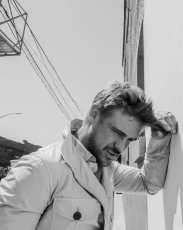 Monochrome portrait of adult male in a collared jacket, head tilted, hand in hair, with an urban background of building and utility lines.