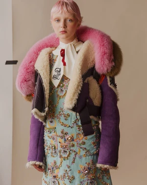 Woman with pink hair wears a blue patterned dress and a pink-trimmed jacket in a studio portrait. Fashion and beauty editorial photography.
