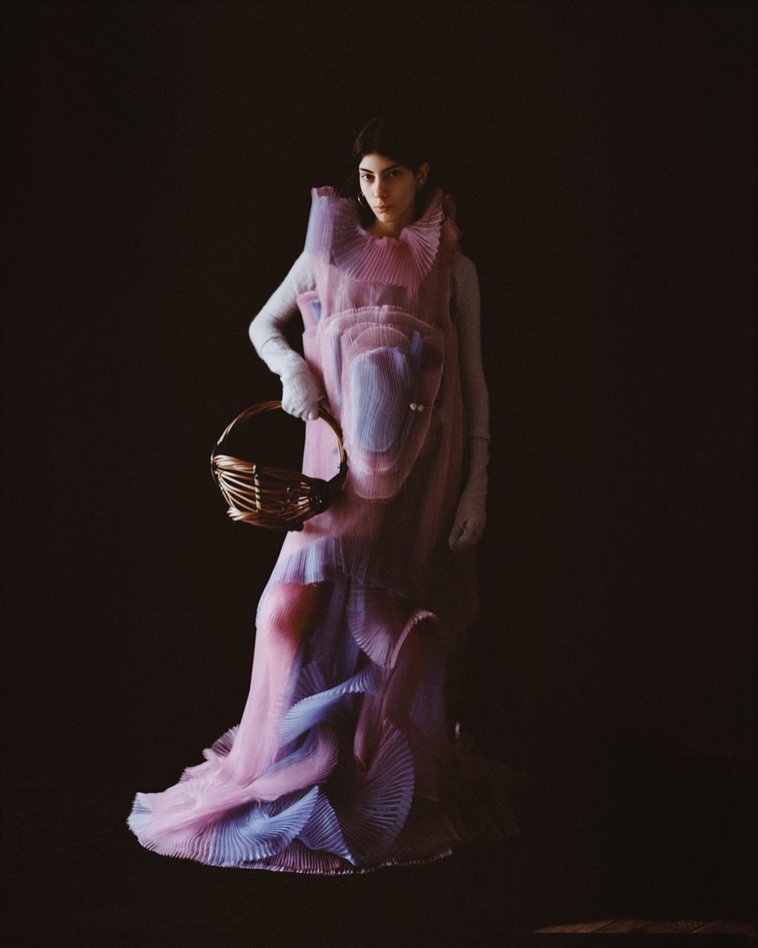 A woman stands in a pink and blue dress, holding a woven basket, in a studio setting with a dark background in this fashion shot.