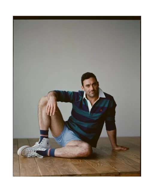 A man sits on a wood floor in a blue and green striped collared shirt, blue shorts, white socks, and sneakers with a white wall background.