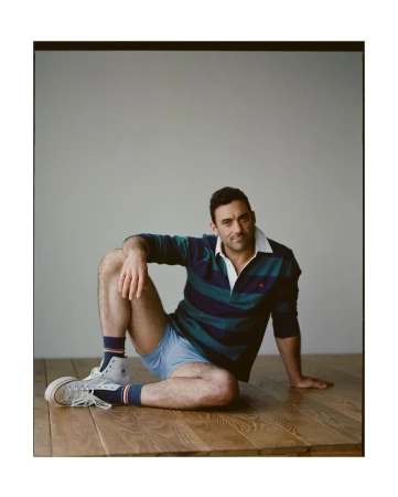 A man sits on a wood floor in a blue and green striped collared shirt, blue shorts, white socks, and sneakers with a white wall background.